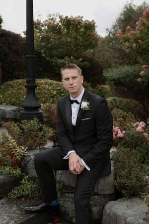 Groom in classic tuxedo poses in garden setting with pink flowering bushes.