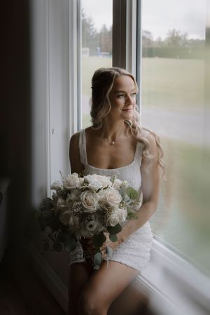 Bride in white dress sits by window holding elegant bouquet of white flowers in soft natural light.