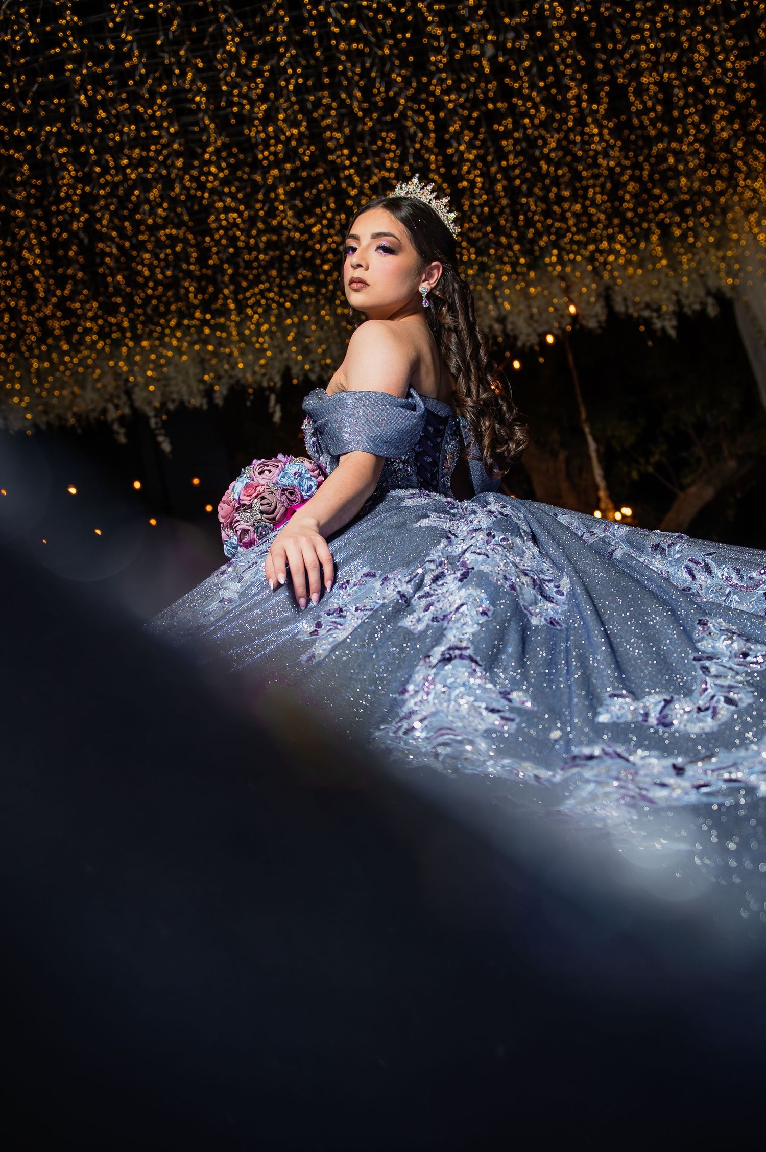 A dramatic portrait of someone in a lavender ball gown against a backdrop of glowing golden fairy lights at night.