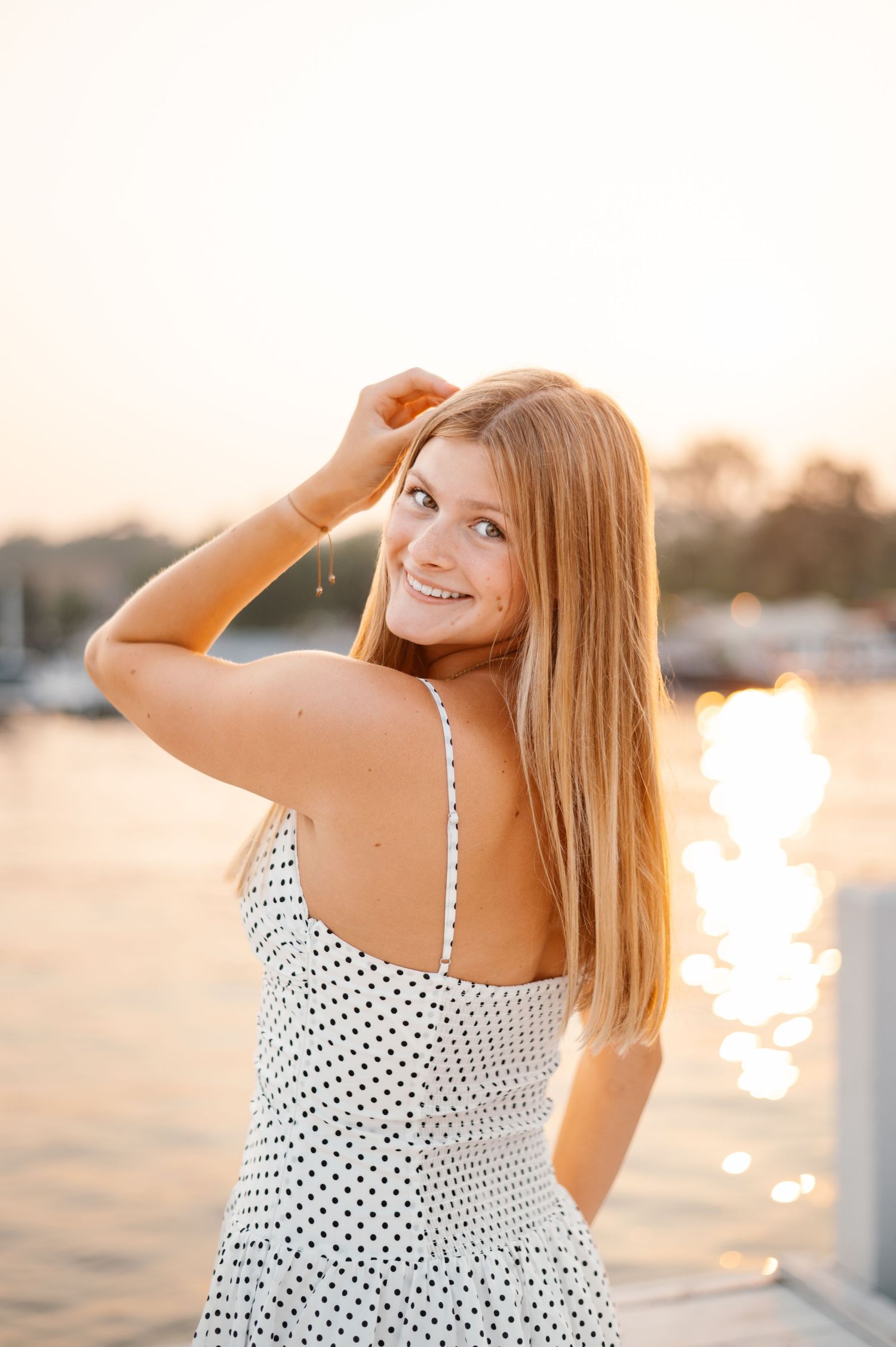 Young woman in white polka dot dress smiling over shoulder at golden hour by waterfront.
