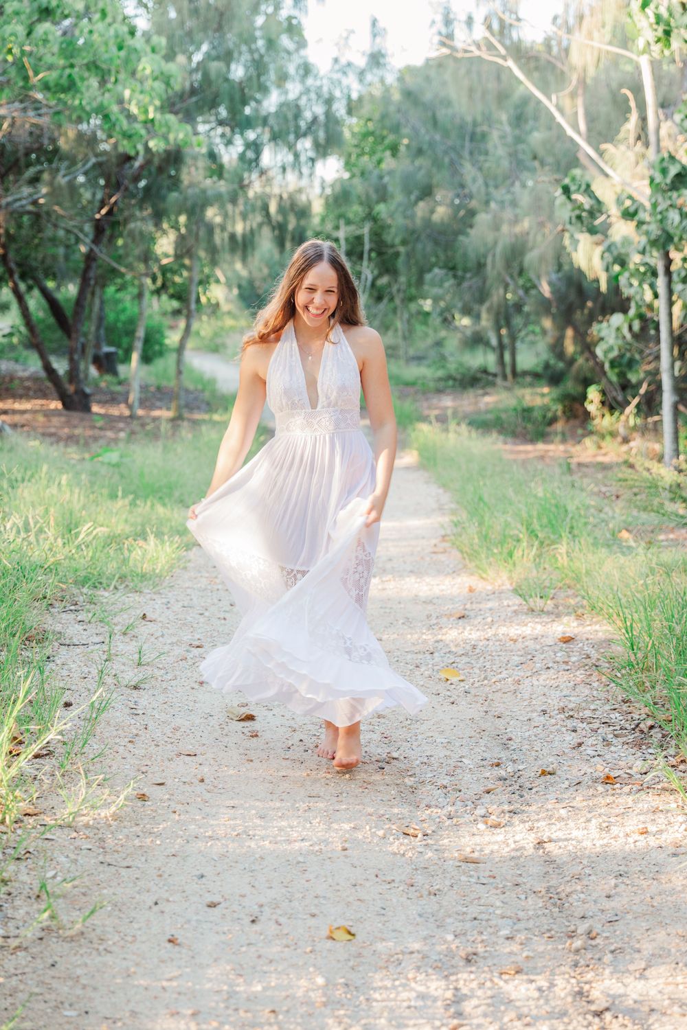 Woman in flowing white halter dress twirling on a sunlit garden path surrounded by lush green trees.