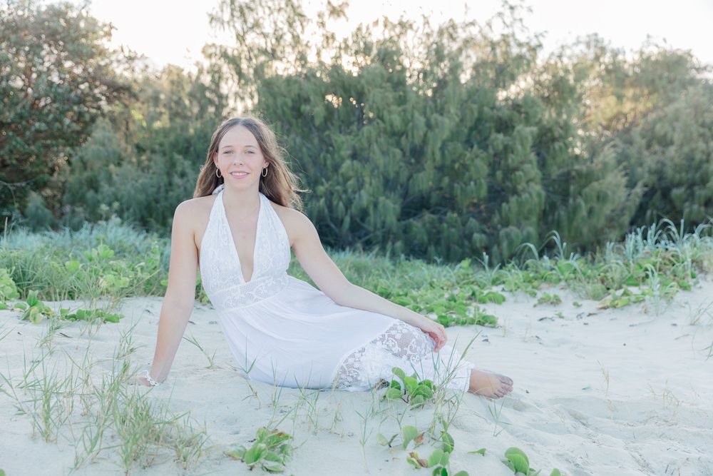 Young woman in white halter dress sitting on sandy beach with lush green trees in soft natural light.