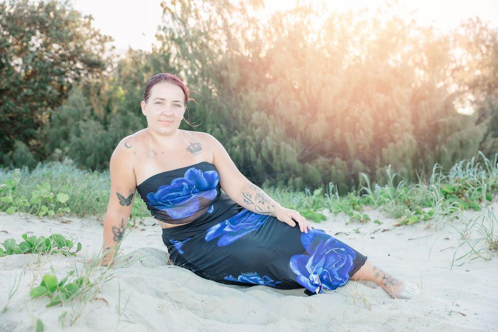 Woman with tattoos in blue floral two-piece outfit sitting on sandy beach with sunlit trees in background.