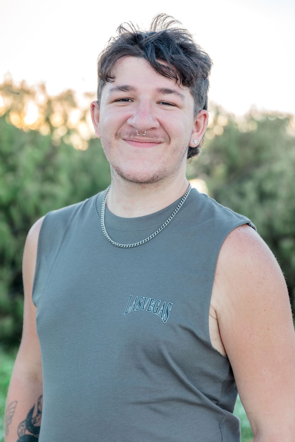 Young man with nose ring and chain necklace smiling outdoors in olive sleeveless shirt with green trees behind.