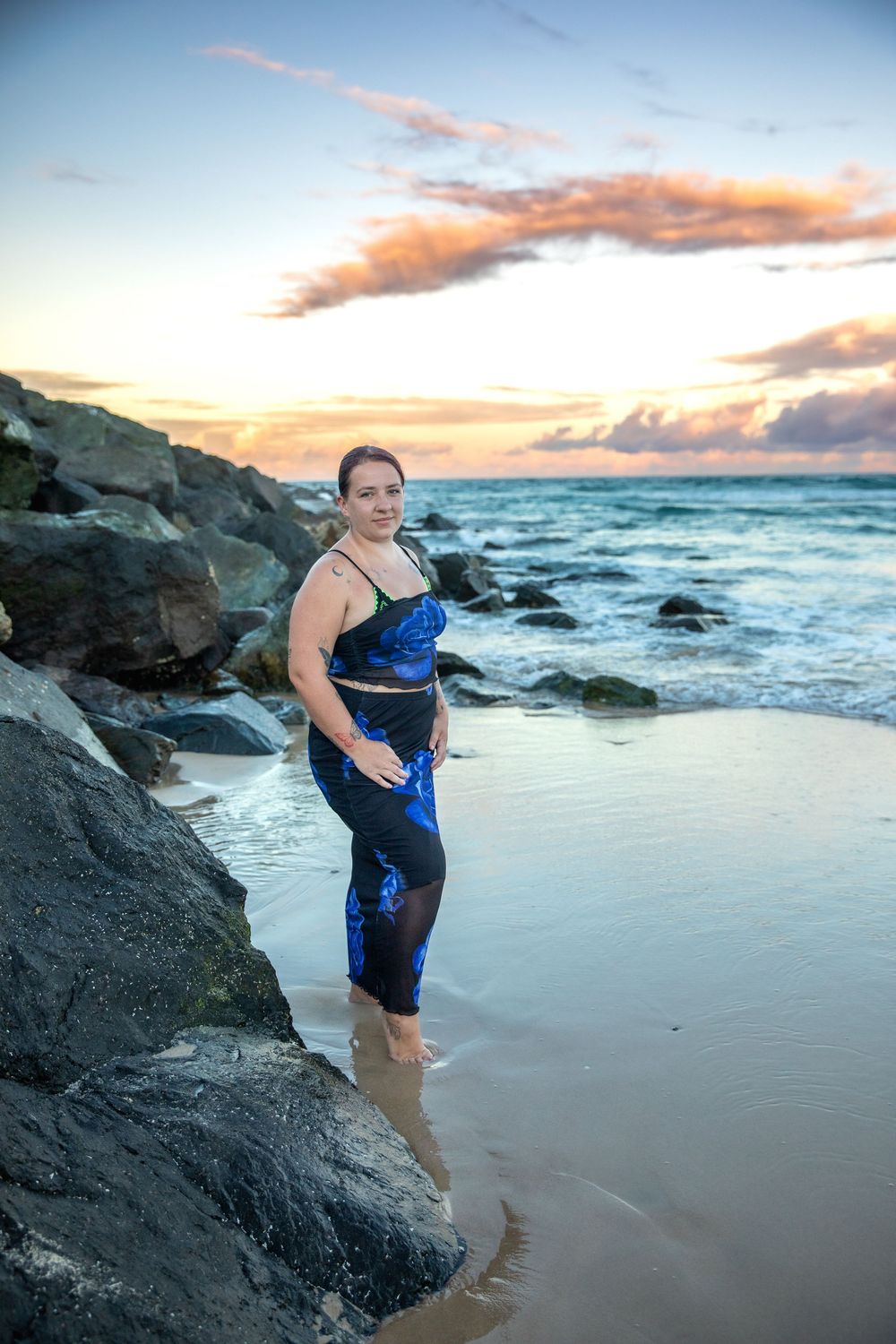 Woman in blue sequin outfit standing on sandy beach near rocks at sunset with dramatic cloudy sky.