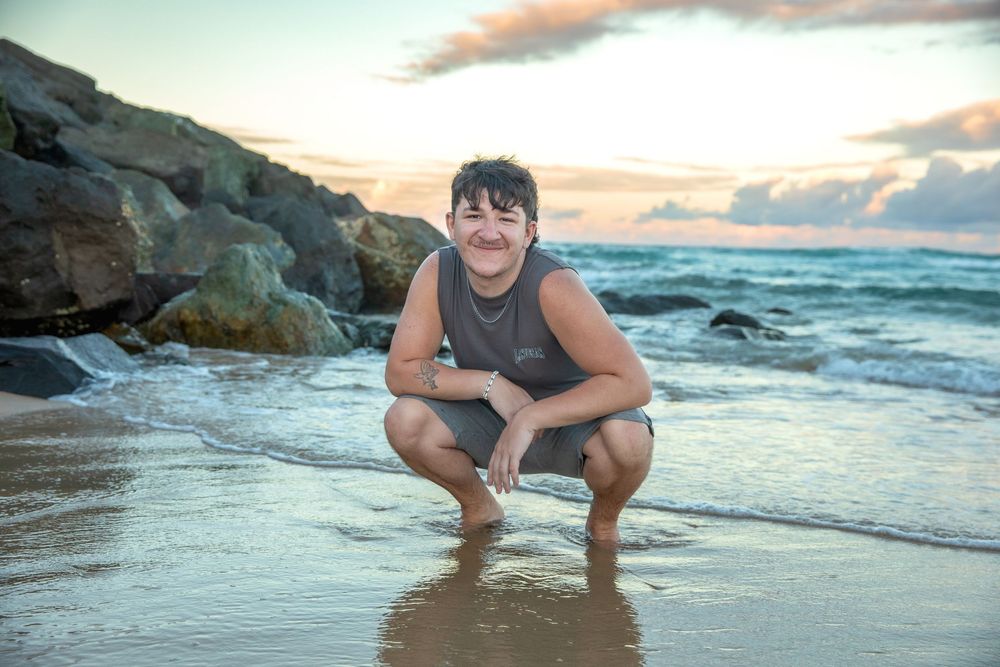 Young person crouching on sandy beach at sunset, smiling, with ocean waves and rocky cliffs in background.