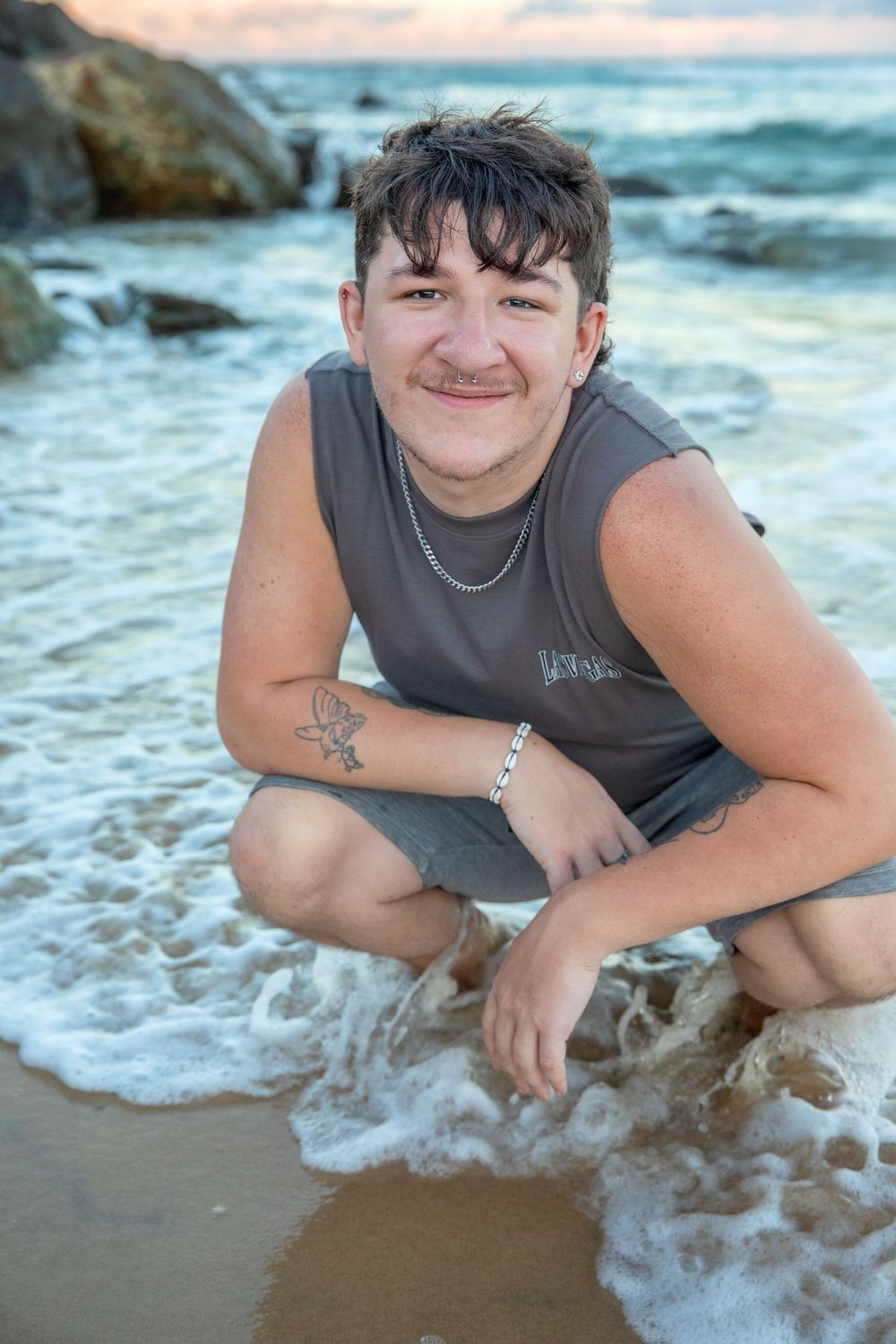 Young person crouching at beach shoreline, smiling, wearing gray tank top with tattoos and jewelry, waves washing over sand.