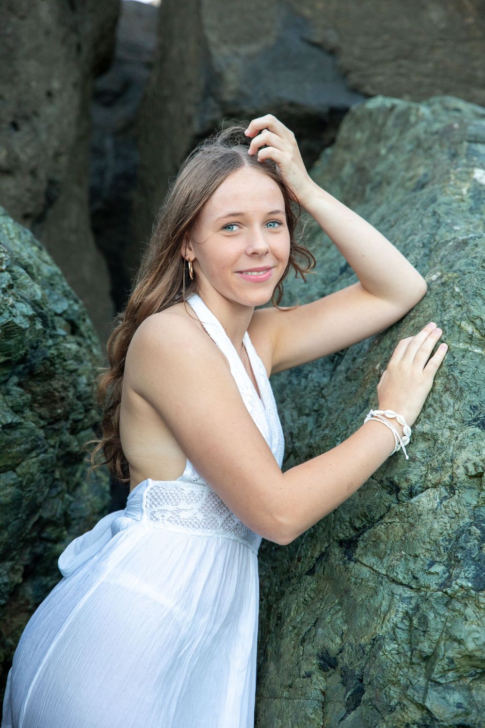 Young woman in white halter dress posing gracefully against large green rocks outdoors.