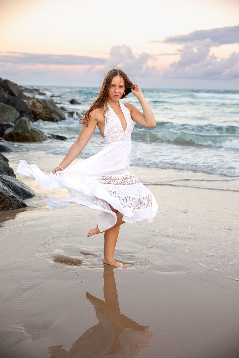 Young woman in white flowing dress dancing barefoot on sandy beach at sunset with ocean waves and rocks.