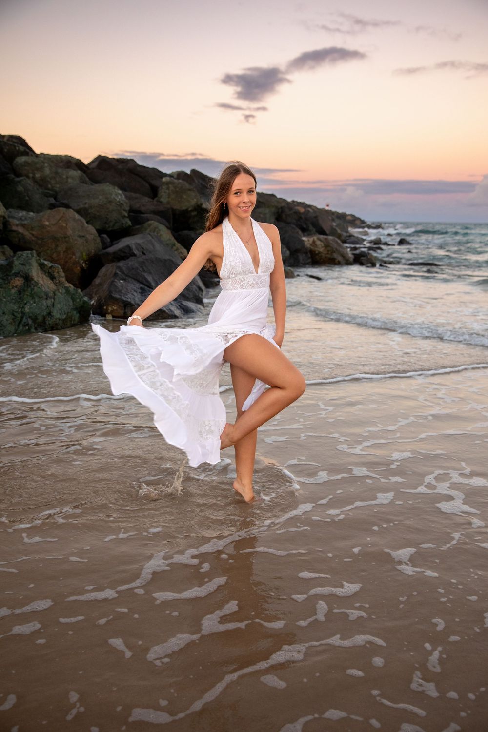 Woman in white flowing dress dancing on beach at sunset with rocky shoreline and ocean waves in background.