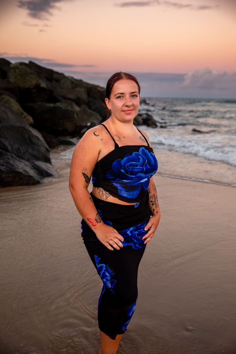 Woman in black and blue floral outfit posing on a sandy beach at sunset with rocky shoreline backdrop.