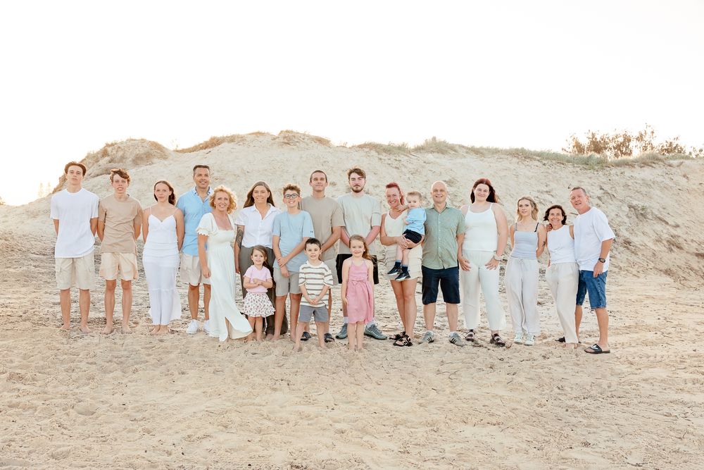Large family group portrait on sandy beach, wearing coordinated white and neutral outfits, with sand dunes in background.