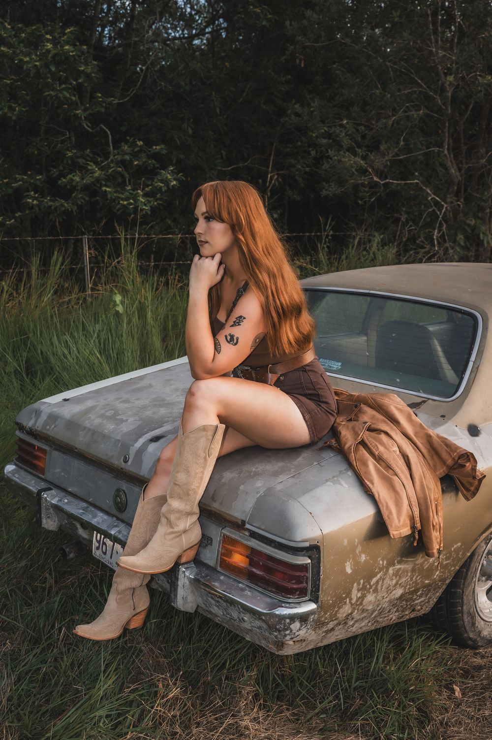 Redhead woman in short dress and knee-high boots sitting on vintage rusty car trunk in rural field.