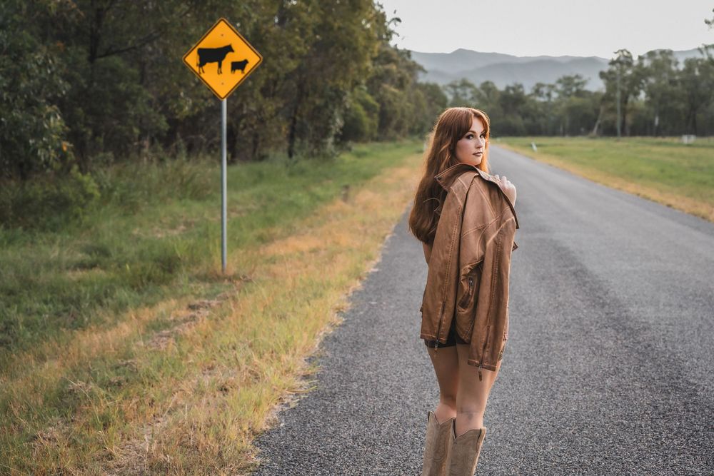 Young woman in brown jacket and boots standing on country road beside yellow cattle warning sign.