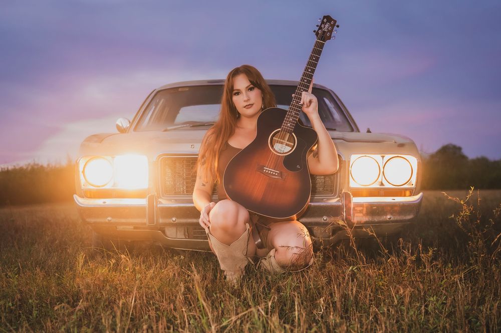 Woman with guitar kneeling before vintage car at sunset in golden field, warm dramatic lighting.