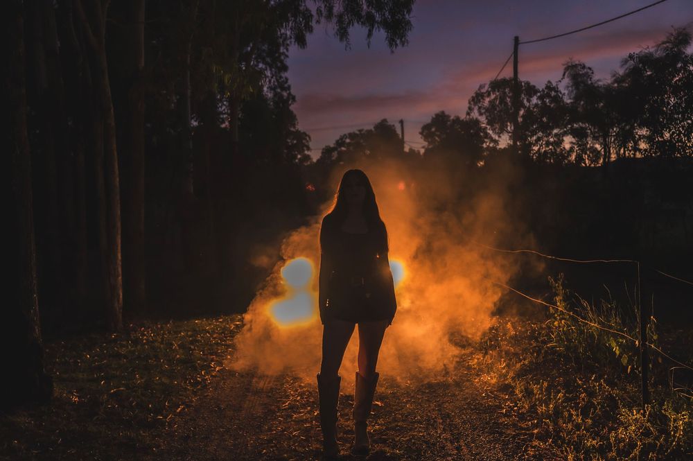 Silhouette of woman standing in dark forest path surrounded by dramatic orange smoke at night.