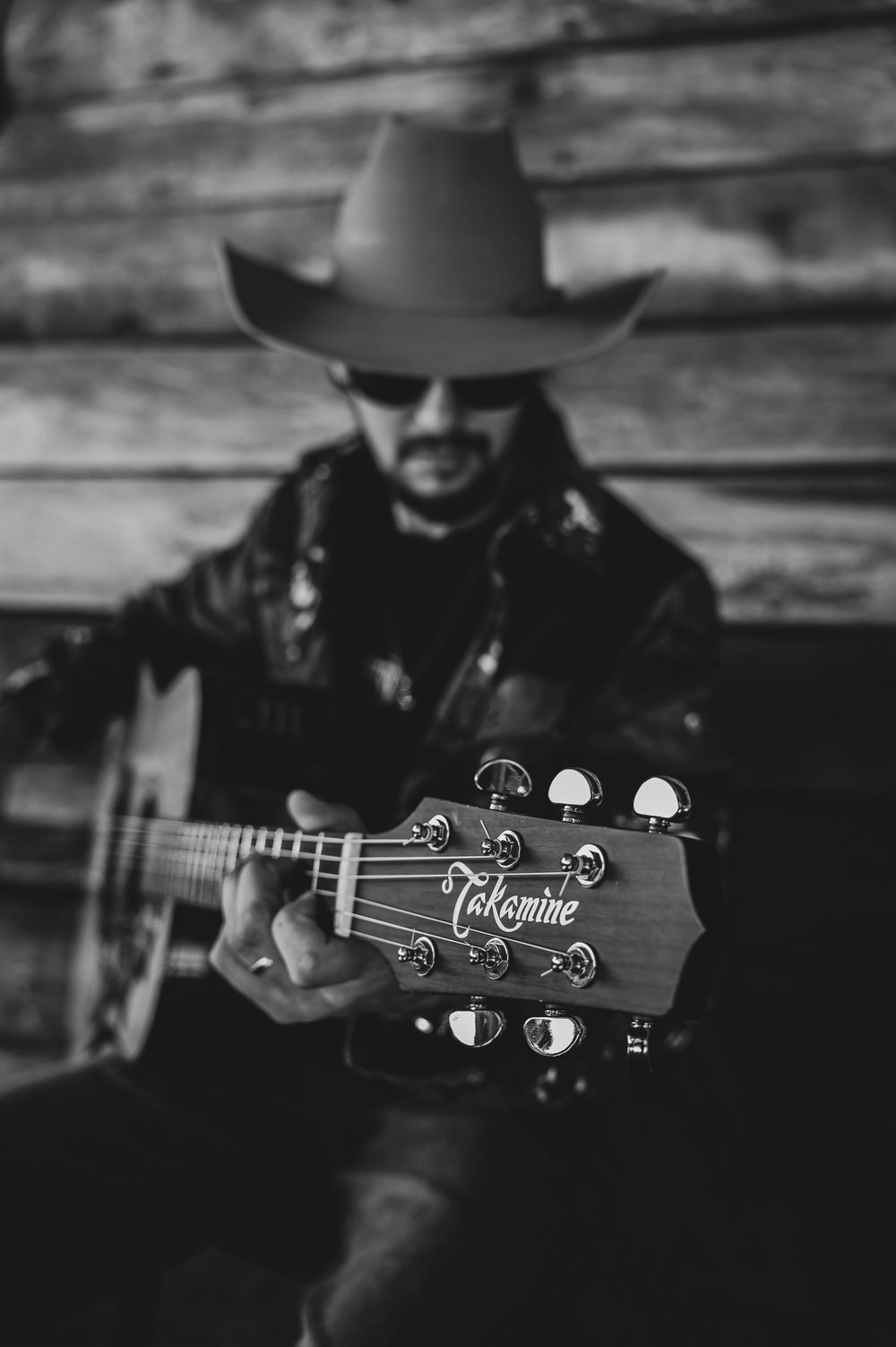 Black and white photo of a man in cowboy hat holding a Takamine acoustic guitar toward camera in rustic setting.