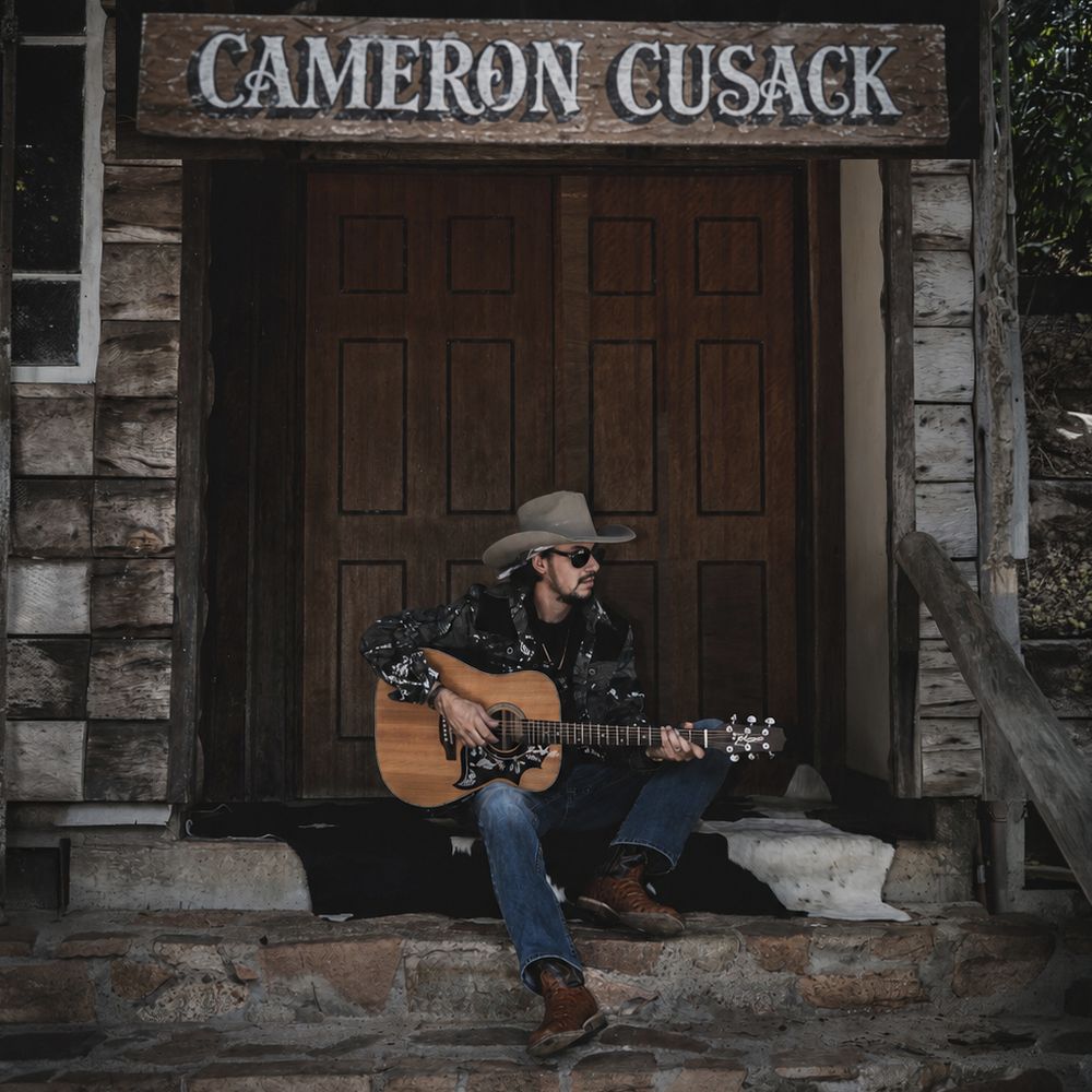 Cameron Cusack sits on stone steps playing acoustic guitar under his name sign on a rustic wooden building.