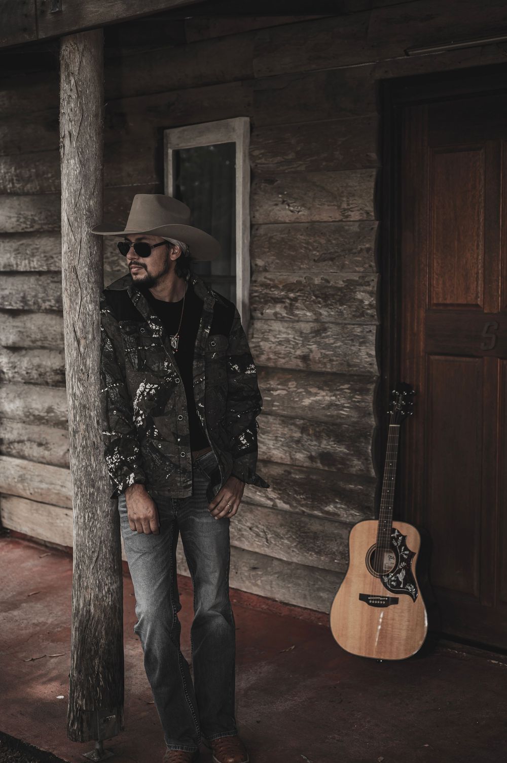 Country musician in cowboy hat and sunglasses leaning against log cabin post with acoustic guitar nearby.