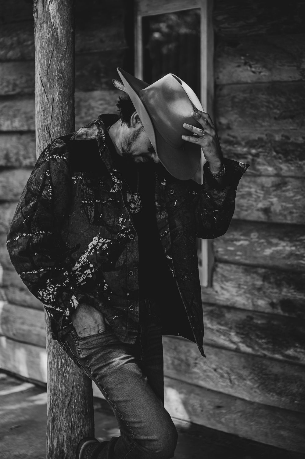 A man in a cowboy hat and embroidered jacket leans against a wooden post outside a rustic log cabin.