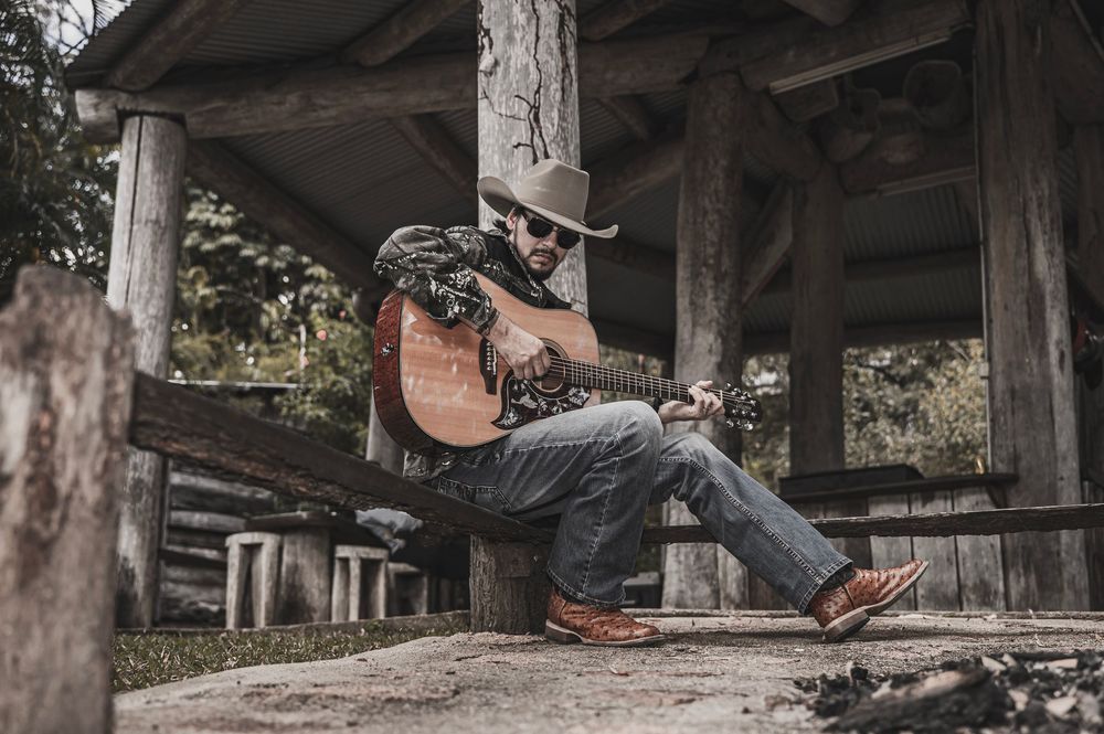 Country guitarist in cowboy hat playing acoustic guitar outdoors under rustic wooden shelter.