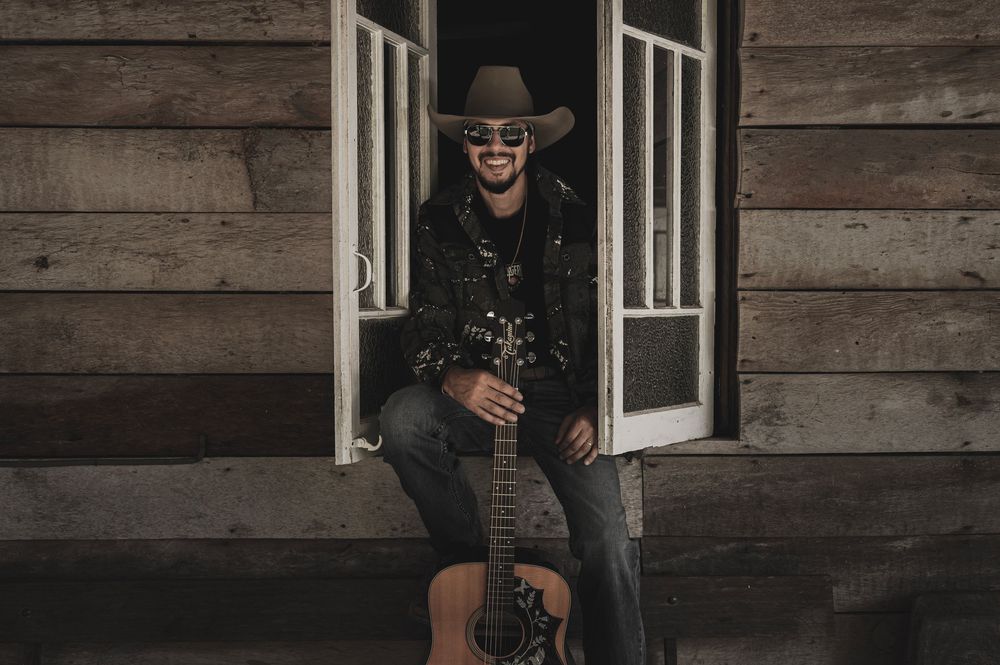 Smiling male country musician in cowboy hat holding acoustic guitar, seated in rustic wooden cabin doorway.