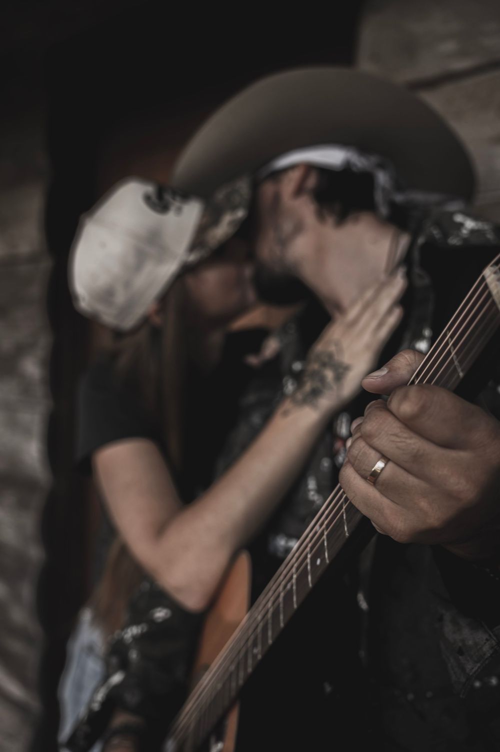 A tattooed guitarist wearing a cowboy hat shares a kiss while playing an acoustic guitar, ring visible on finger.