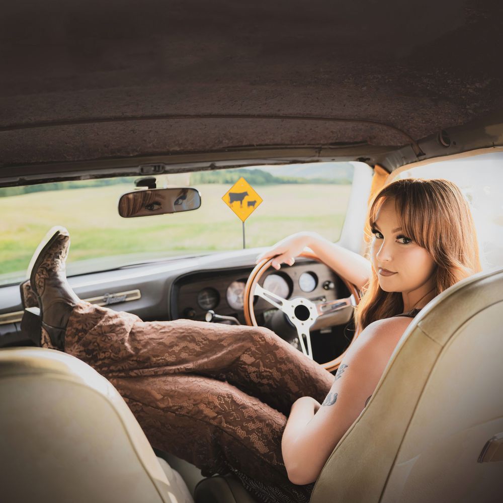 Young woman with cowboy boots on dashboard of vintage car, leaning on steering wheel near rural road with cattle sign.