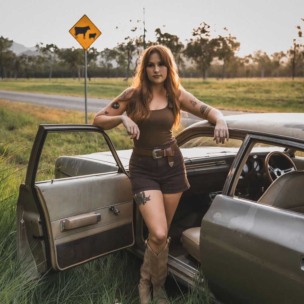 Woman in brown outfit leaning against vintage car on rural road with cattle warning sign in background.