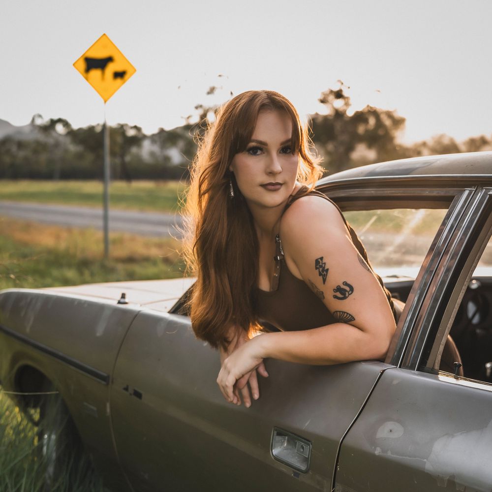 Red-haired woman with tattoos leaning out of vintage car near cattle crossing sign at sunset.