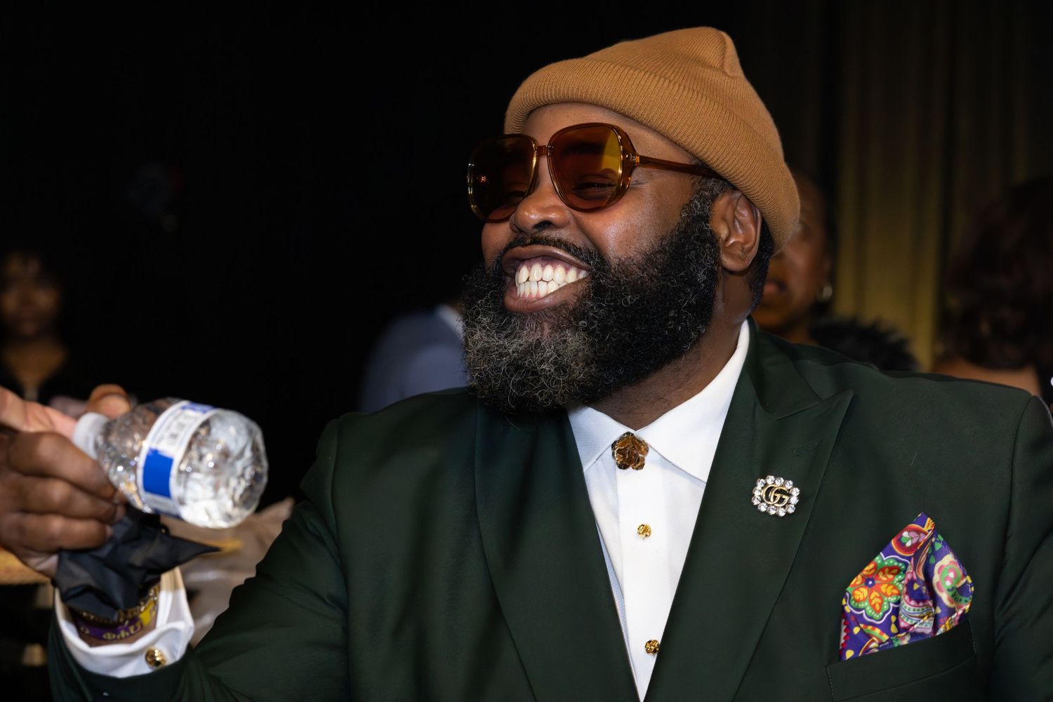 A well-dressed person in a green blazer and beanie smiles while holding a drink at an event.