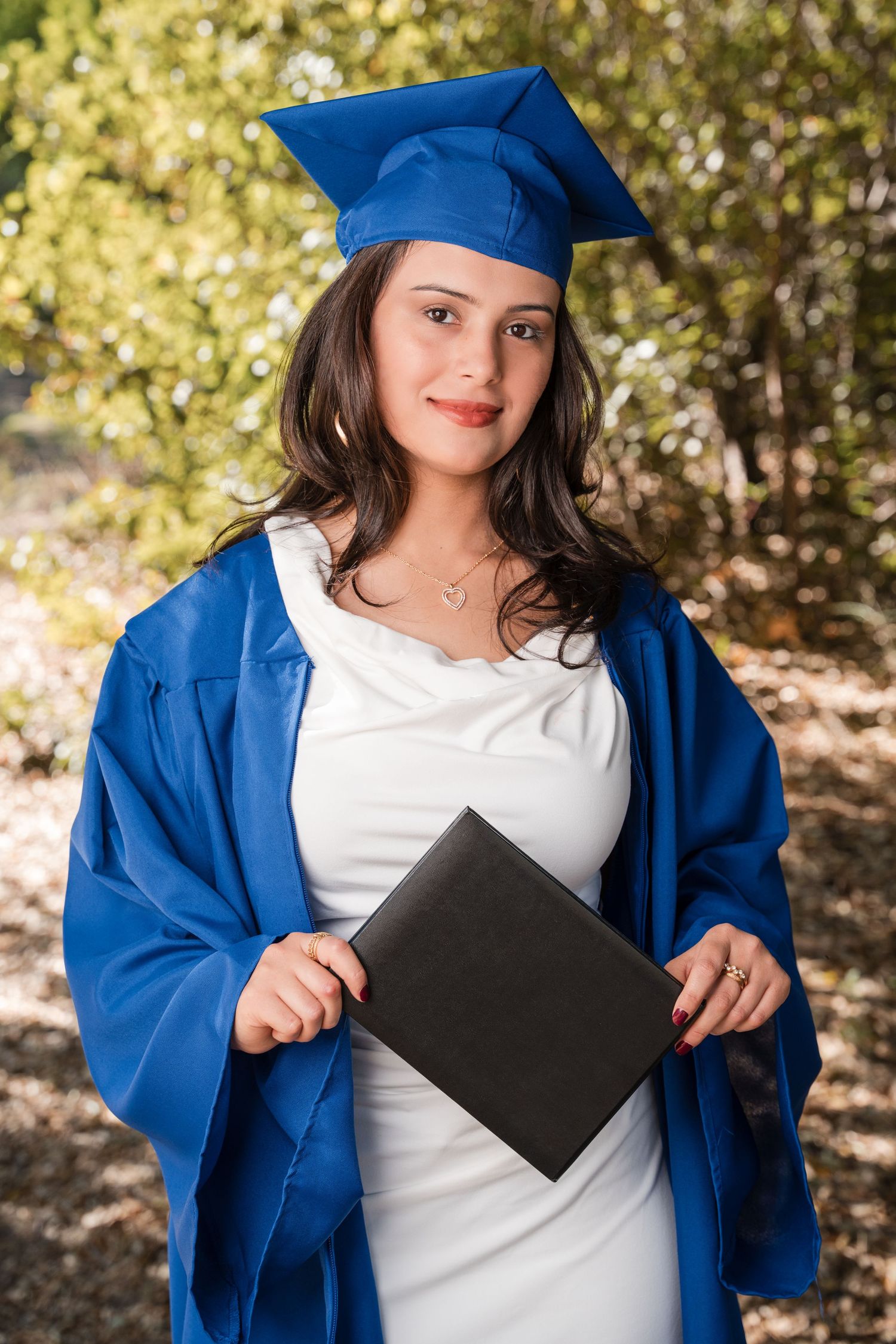 Graduate in bright blue cap and gown holds diploma folder while posing outdoors on sunny day.
