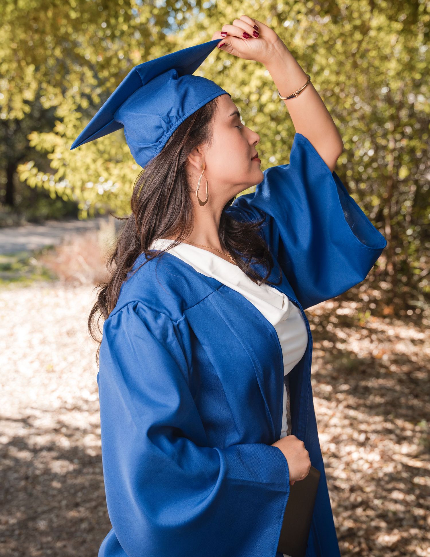 Graduate in royal blue academic regalia adjusts cap while standing in autumn outdoor setting.