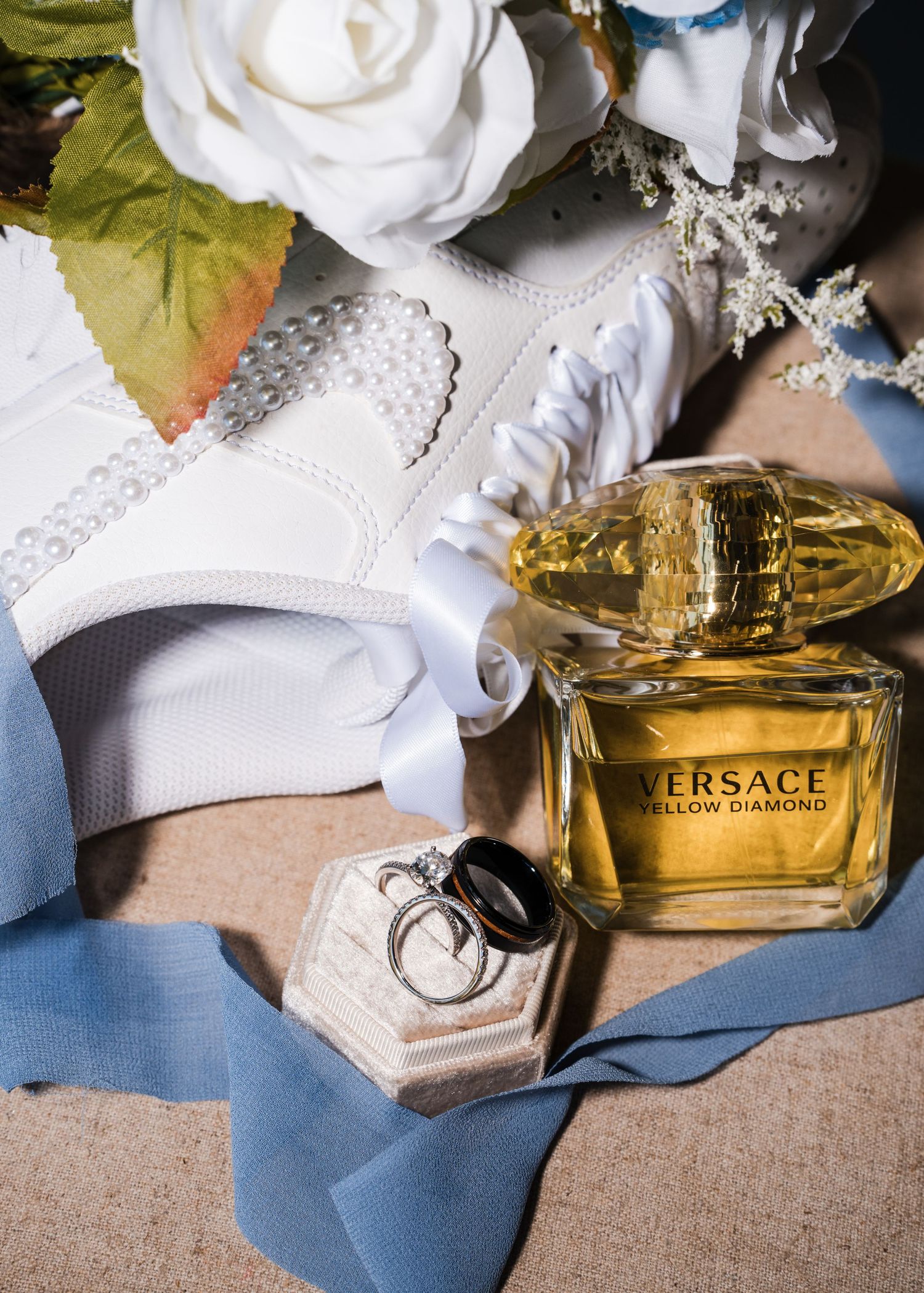 A white rose, Versace perfume bottle, and wedding rings arranged on blue fabric with soft lighting.
