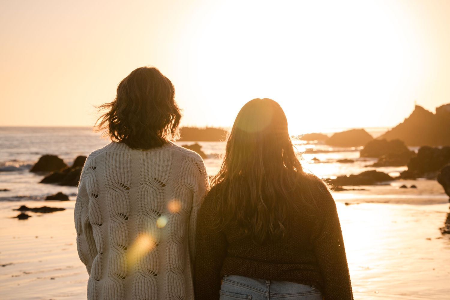 Two people watching sunset together on a rocky beach with ocean waves and golden light filtering through.