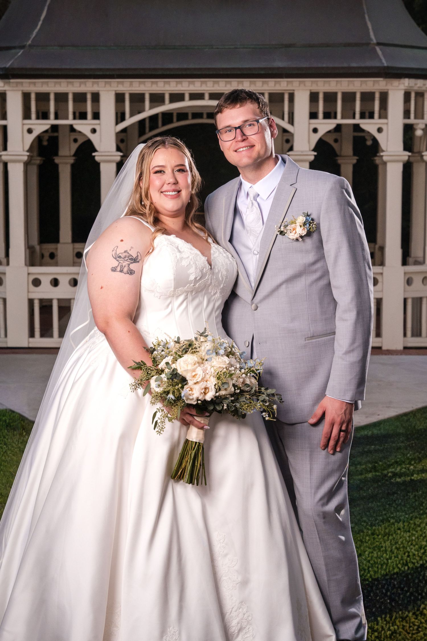 A couple in wedding attire poses together in front of a white gazebo during their outdoor ceremony.