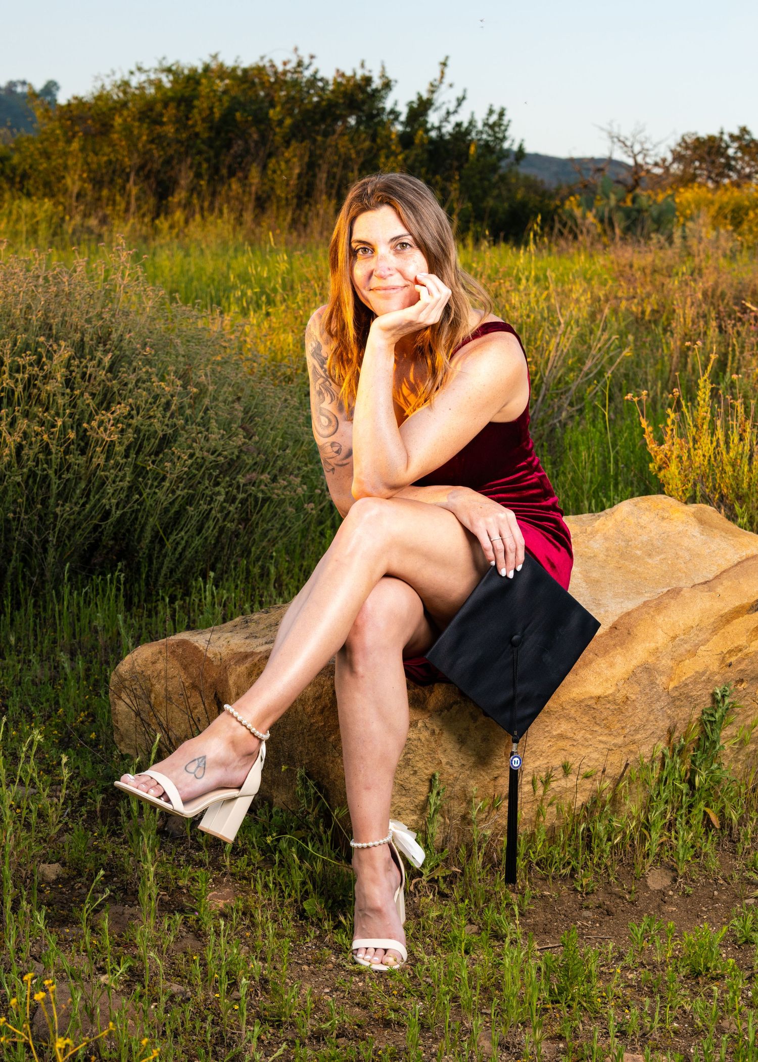 A person in a burgundy dress sits on a large rock during golden hour in a summer field with tall grasses.