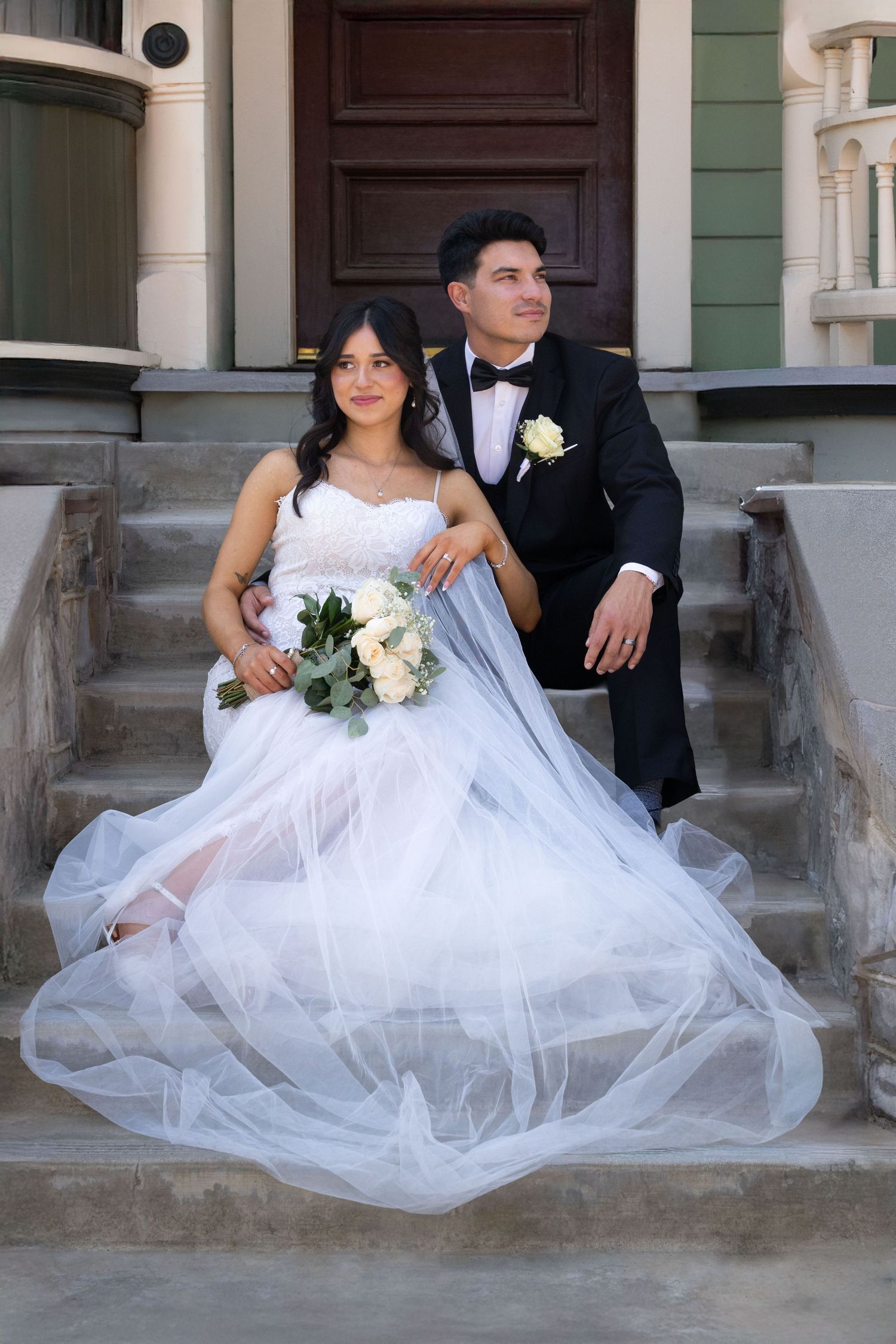 Elegant couple in wedding attire pose together on stone steps, with bride in flowing white gown holding floral bouquet.