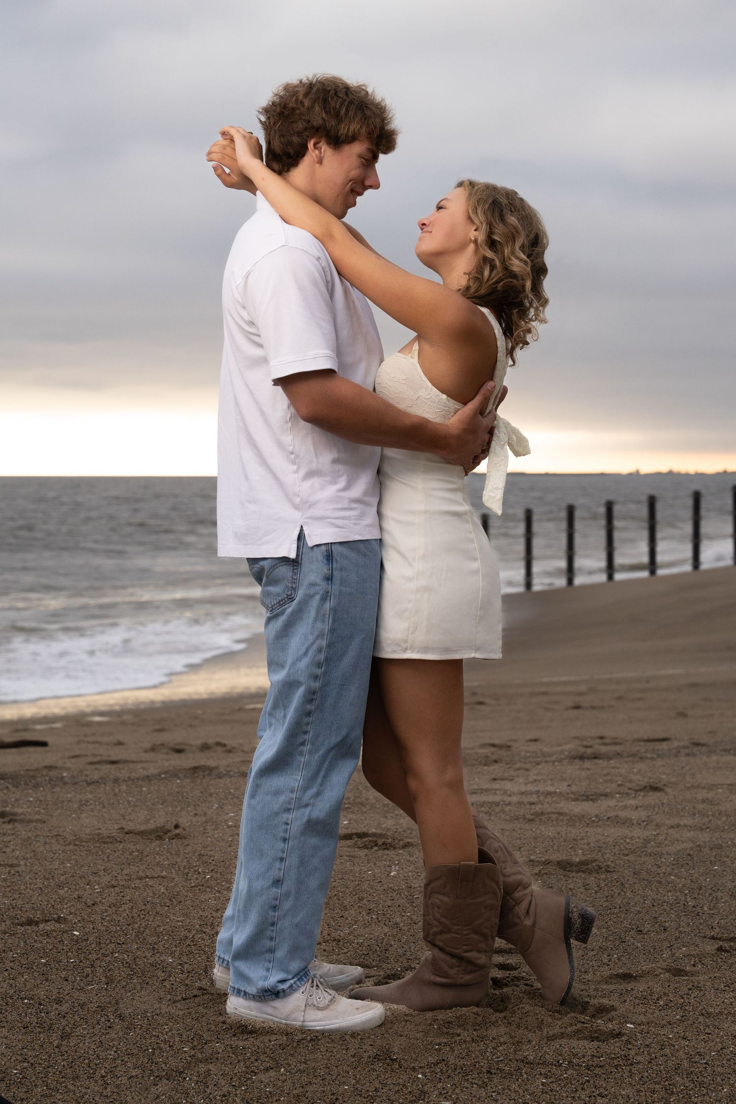 A romantic couple embraces on a cloudy beach at sunset with ocean and pier in the background.