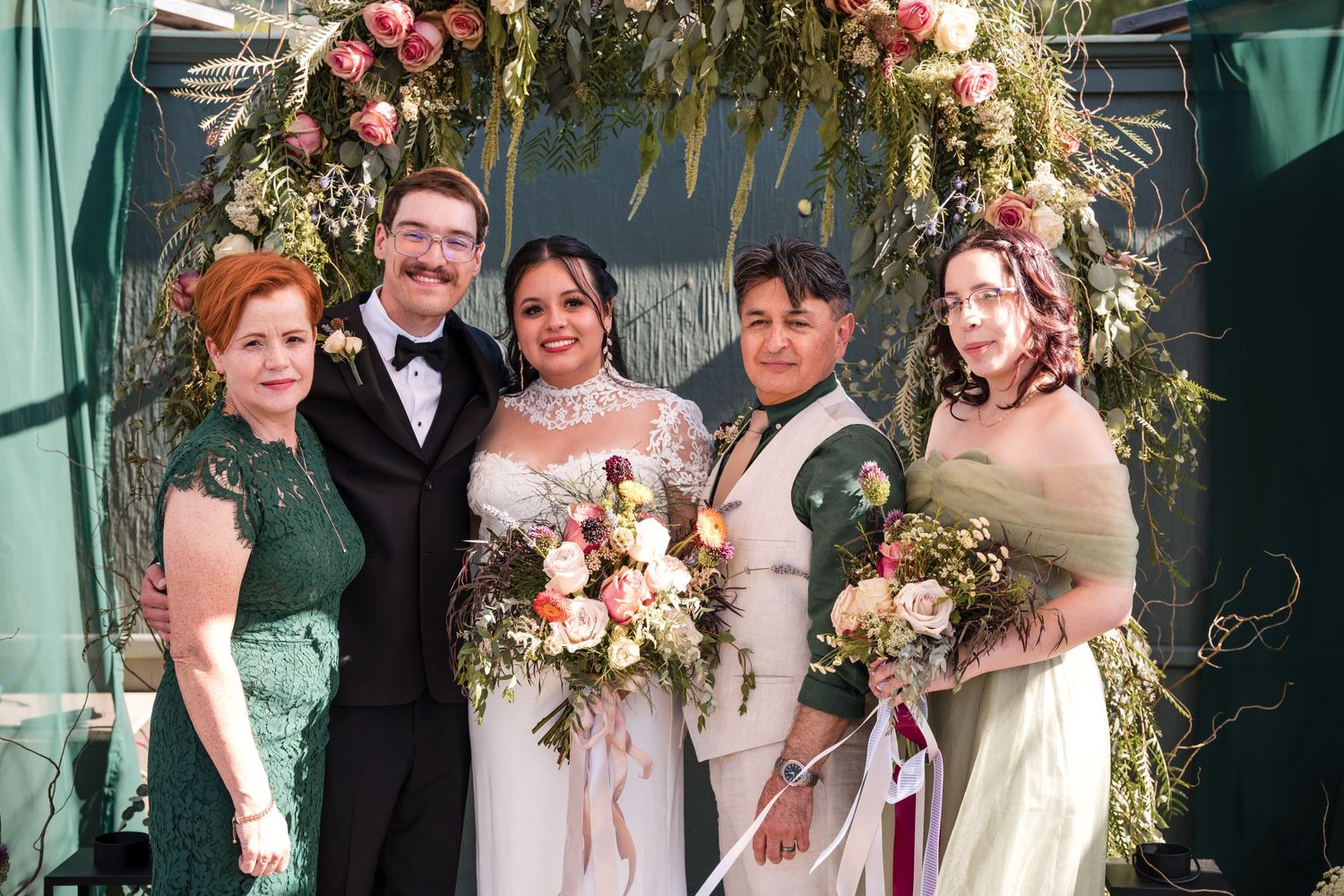 Wedding party posing under floral arch with greenery and pink flowers.