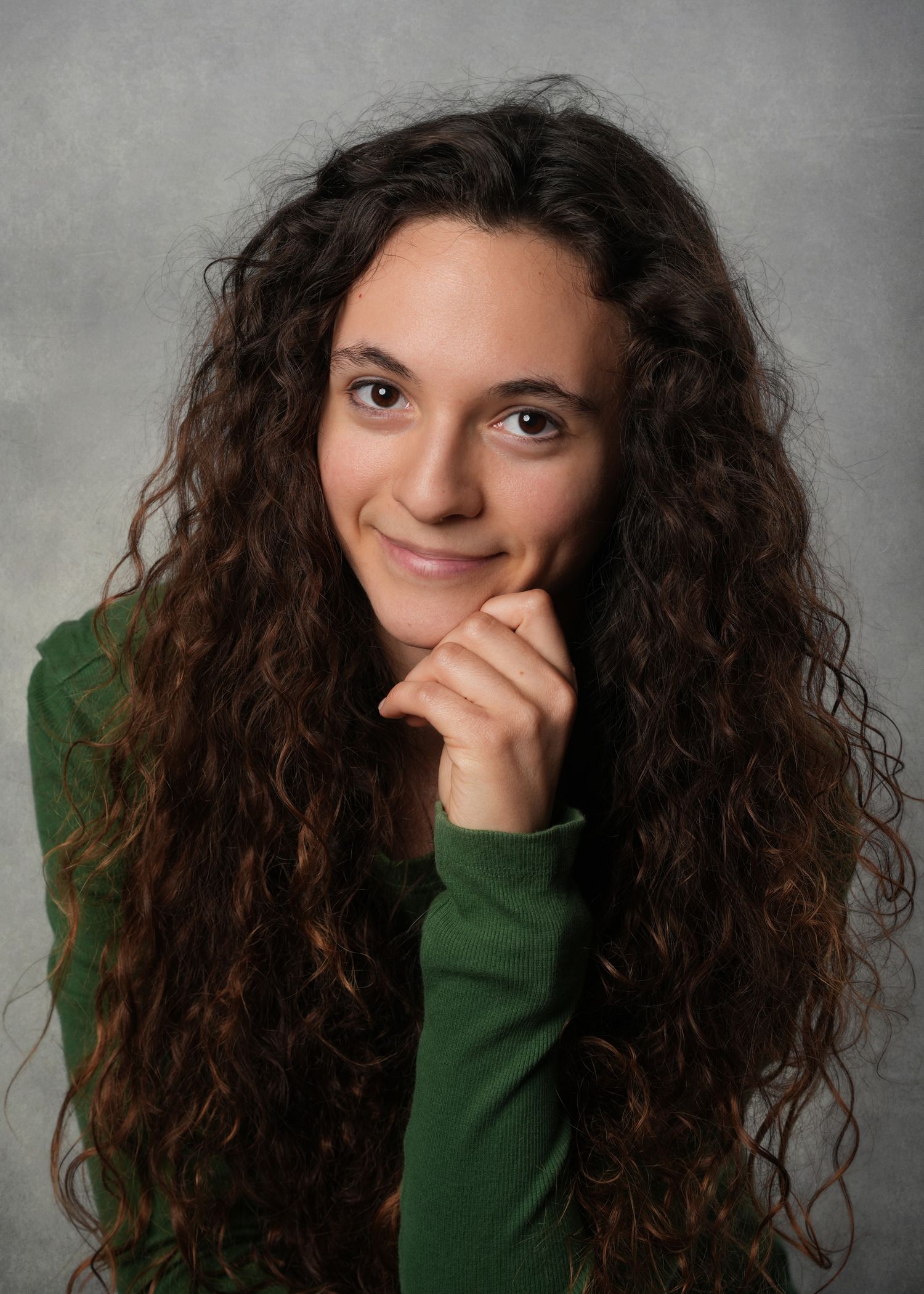 Young woman with long curly brown hair wearing a green turtleneck, smiling with hand on chin against gray background.