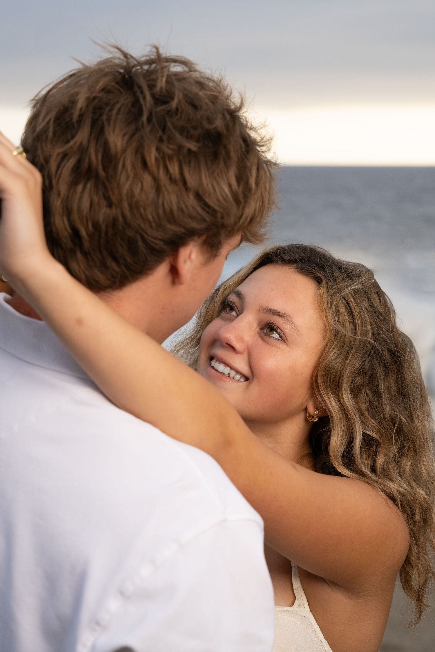 A romantic couple embraces at sunset by the ocean, with the woman smiling warmly at her partner.