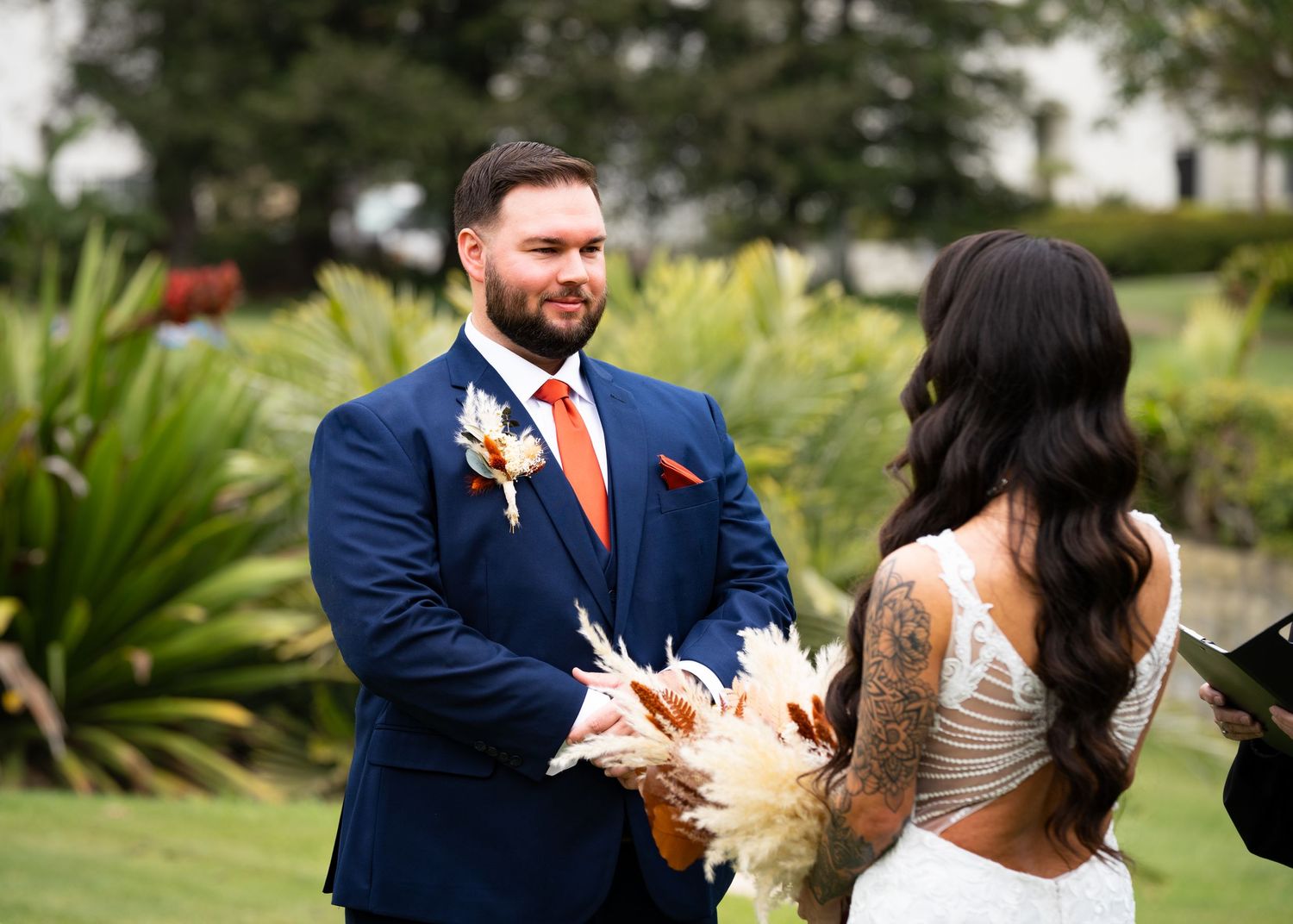 Couple stands together at outdoor garden wedding ceremony with orange floral accents and tropical greenery in background.