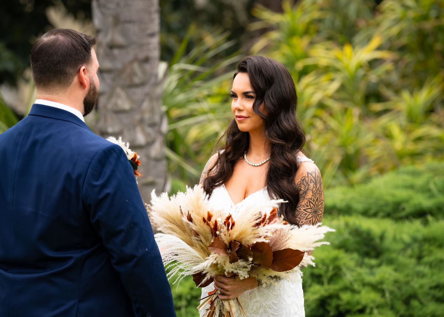 A wedding couple outdoors in a garden setting with dried pampas grass bouquet and lush tropical greenery in the background.