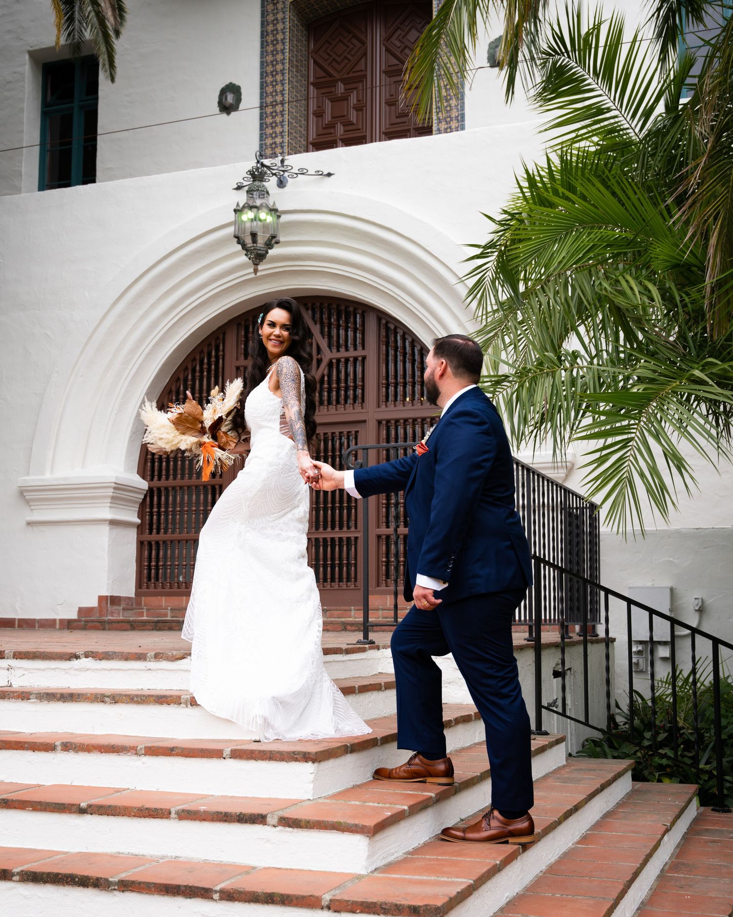 Couple in formal wedding attire ascend white stone steps with palm trees and an arched doorway in the background.