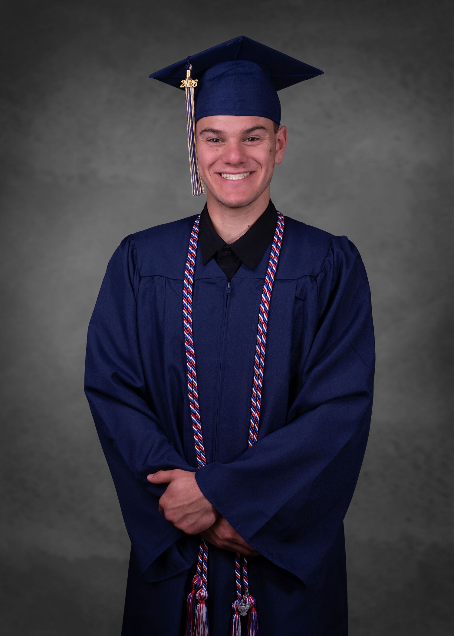 Smiling graduate in navy blue cap and gown with honor cords poses for formal graduation portrait.