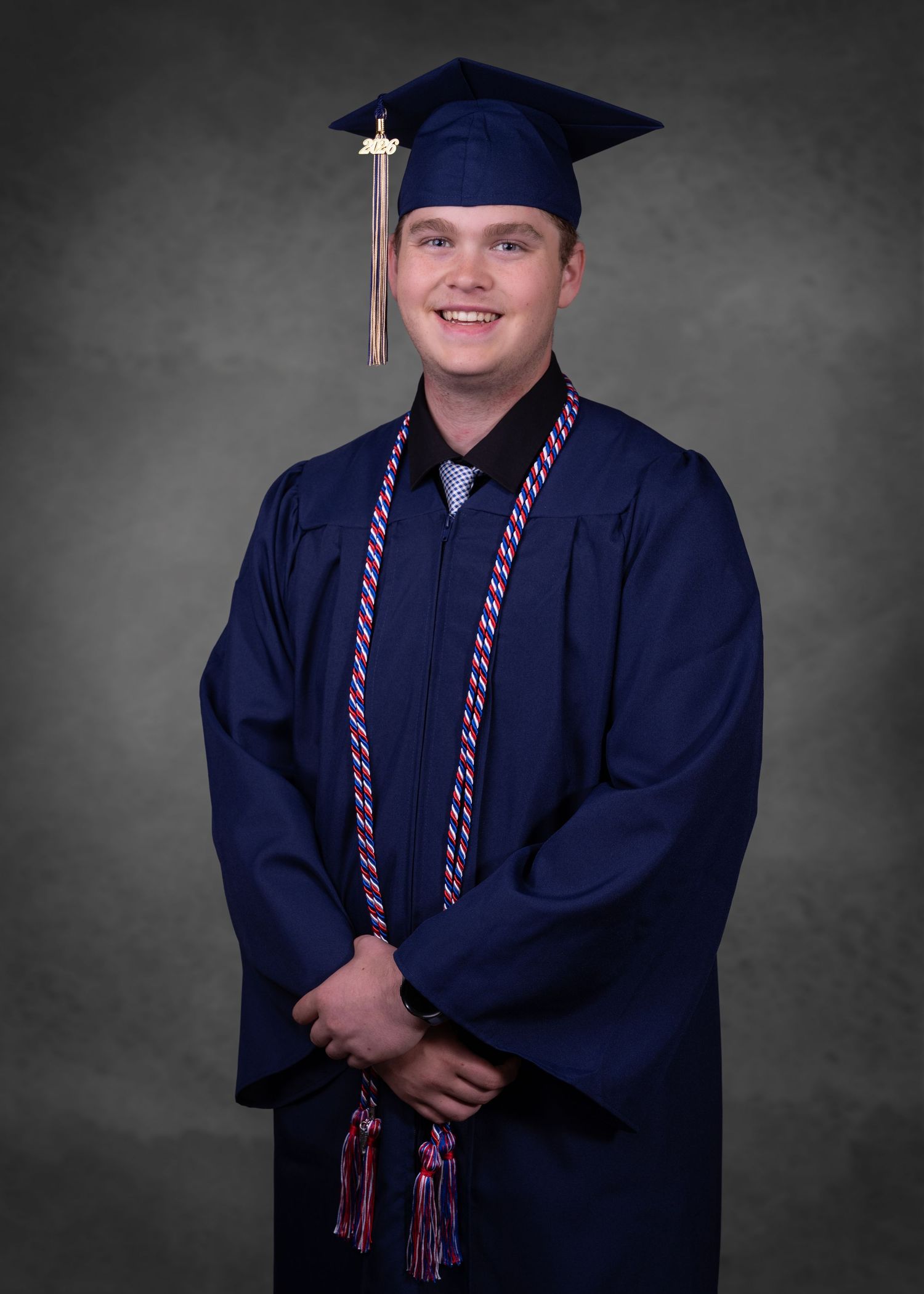 Young man in navy blue graduation cap and gown with honor cords smiles for formal portrait photo.
