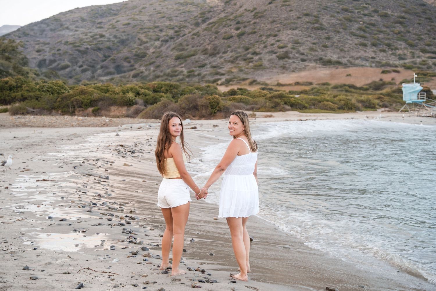 Two people in white dresses walk hand in hand along a scenic Mediterranean beach at sunset.