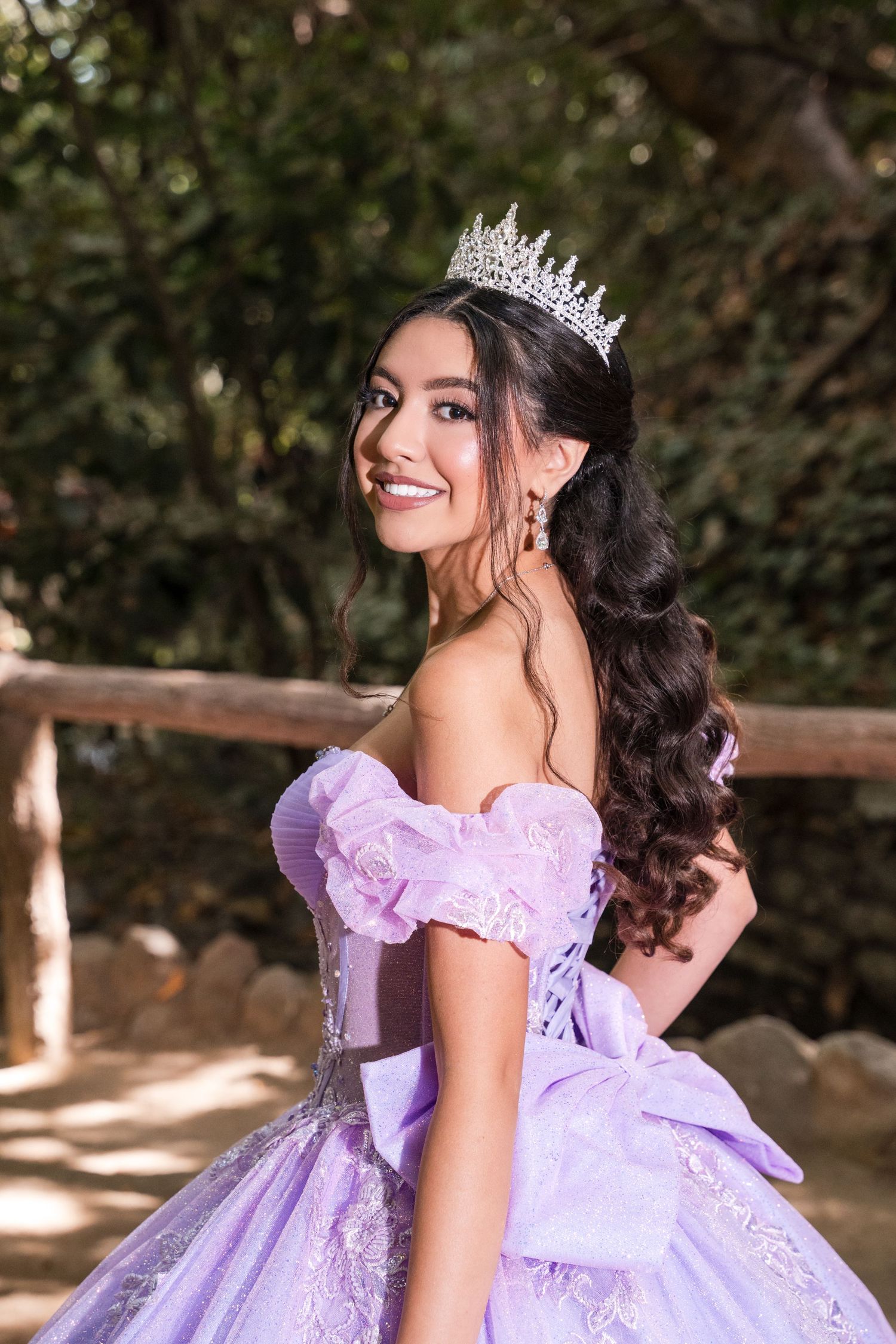 A person wearing a lavender ball gown with puff sleeves and a crown poses gracefully in an outdoor garden setting.