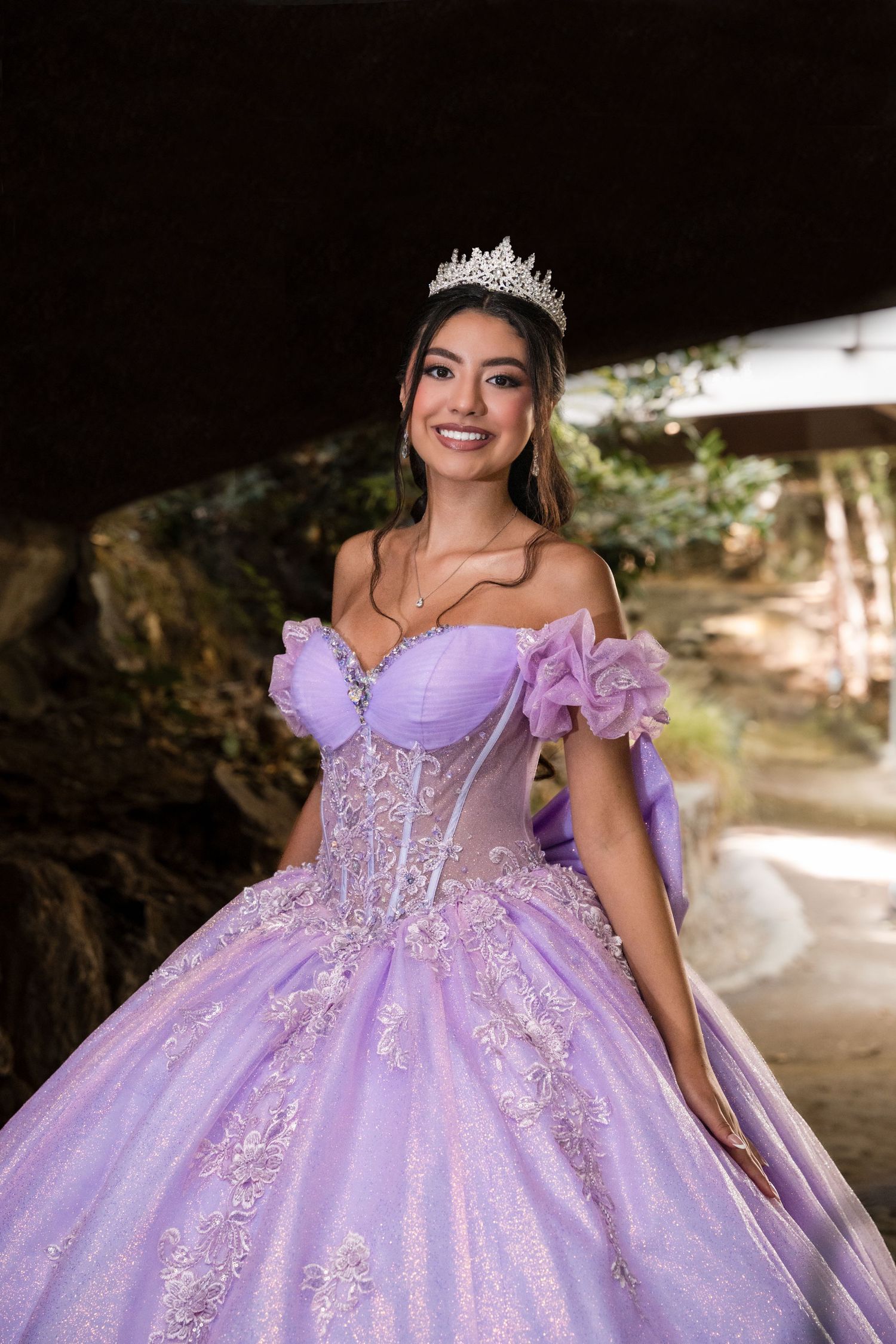 A lavender quinceañera ball gown with off-shoulder design and sparkling crystal embellishments in outdoor setting.